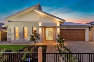 Modern single-storey home extension with twilight lighting, featuring a neutral exterior, wooden garage door, landscaped front yard, and illuminated entrance.