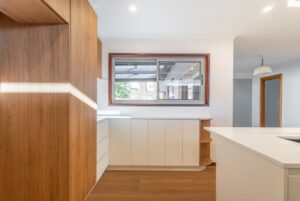 Newly renovated Australian kitchen featuring white cabinetry, wooden accents, a large window overlooking the backyard, and a modern white island bench with integrated storage.