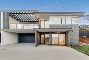 Modern two-storey home with dark brick and white cladding, featuring a spacious driveway, double garage, balcony with glass railing, and large windows