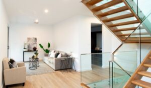 Newly renovated modern living room in an Australian home, featuring a grey sofa, beige armchair, decorative plants, a glass and wood staircase, and light wooden flooring