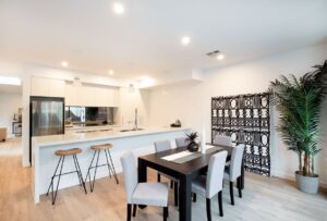 Newly renovated modern Australian kitchen and dining area featuring a long white island with wooden bar stools, a black dining table with grey chairs, light wooden flooring, and decorative plants and artwork