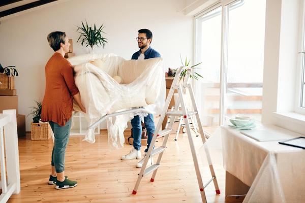 Couple carrying covered furniture while preparing for Home Renovations in a bright living space with ladders and renovation materials nearby